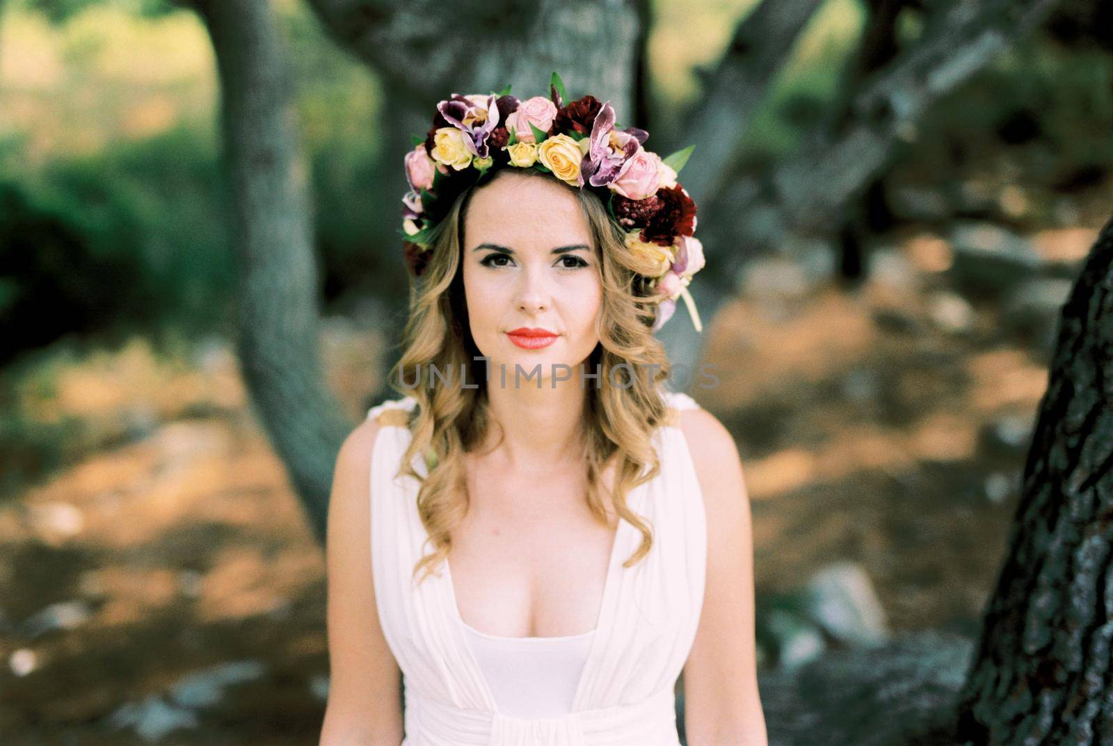 Bride in a wreath of multi-colored flowers on her head stands near a tree. High quality photo