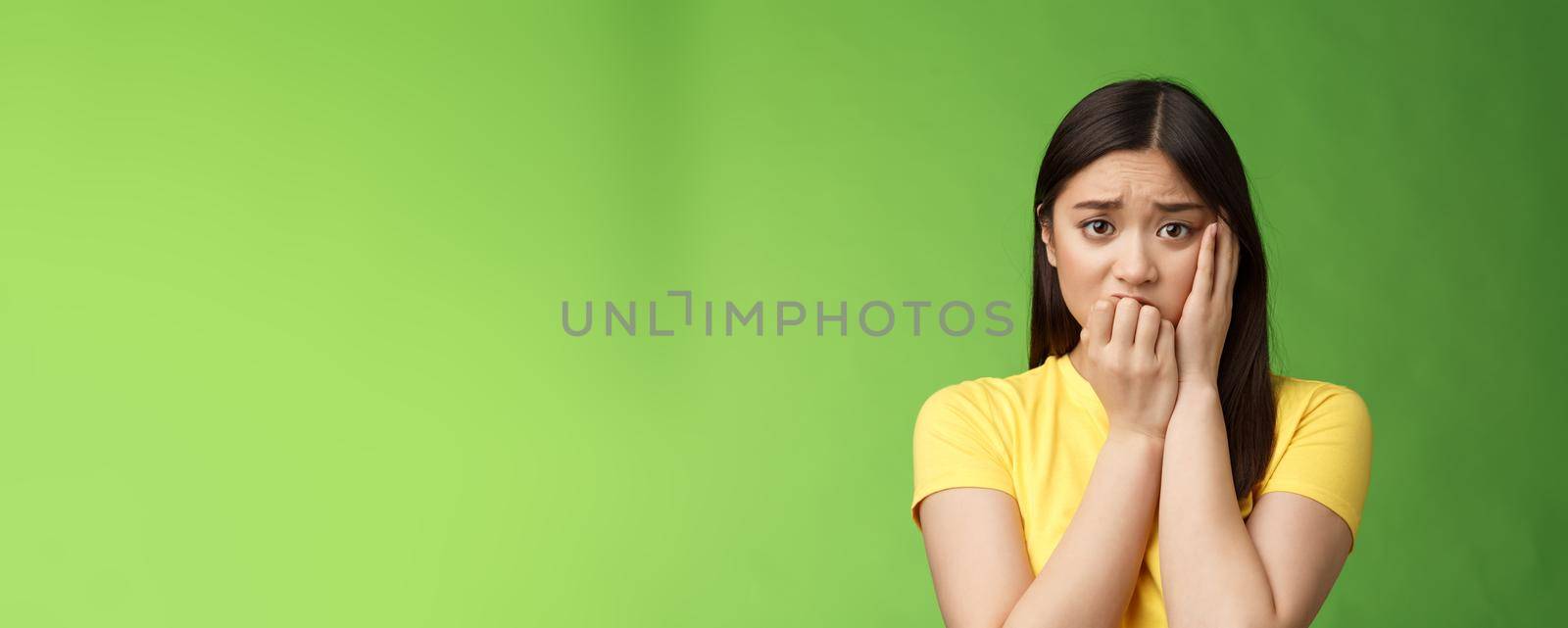 Innocent insecure timid asian scared girl panicking, standing afraid victim terrified, touch cheek shocked, frowning stunned, biting fingernails, anxiously stare camera, green background by Benzoix