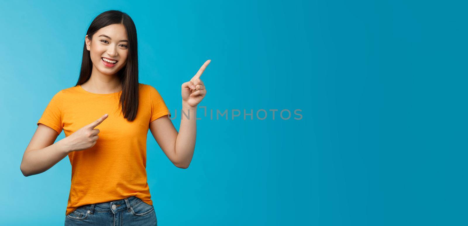 Cheerful lively cute asian woman smiling laughing upbeat, pointing upper right corner, give advice what choose, shopping online, grinning happily, enthusiastic promo, blue background.