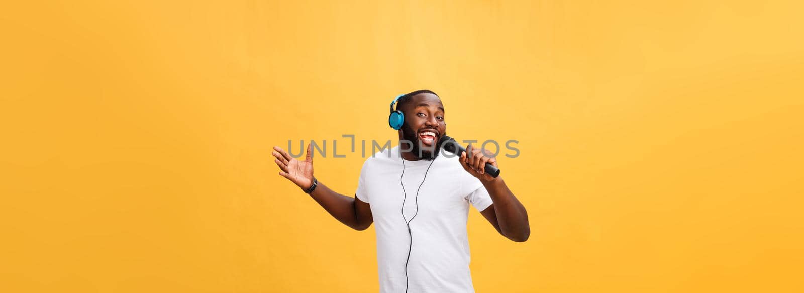 Portrait of cheerful positive chic. handsome african man holding microphone and having headphones on head listening music singing song enjoying weekend vacation isolated on yellow background.