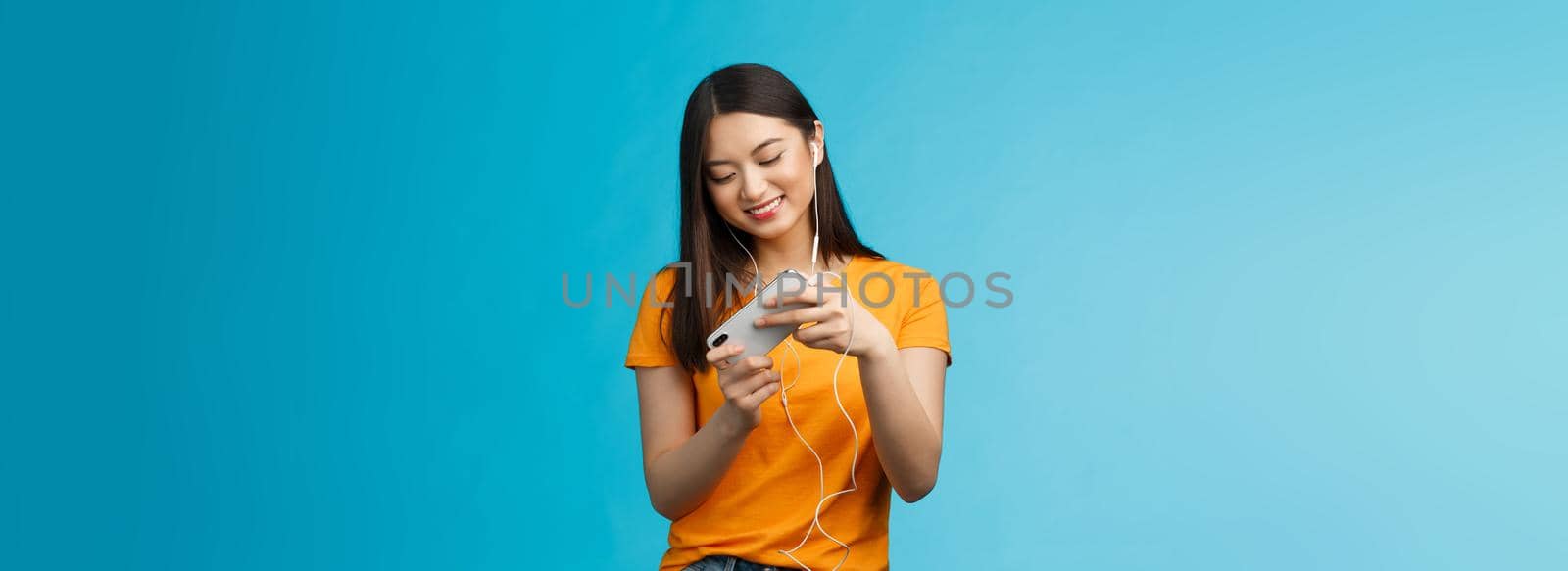 Cute entertained good-looking urban female student spend break playing racing game, hold smartphone horizontal tilt phone move car, smiling broadly standing enthusiastic blue background.