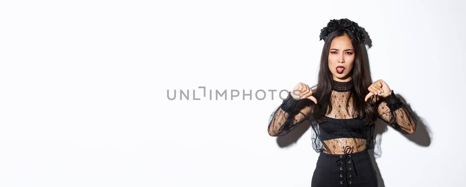 Image of disappointed asian woman wearing halloween costume of witch, showing thumbs-down and sticking tongue displeased, white background.