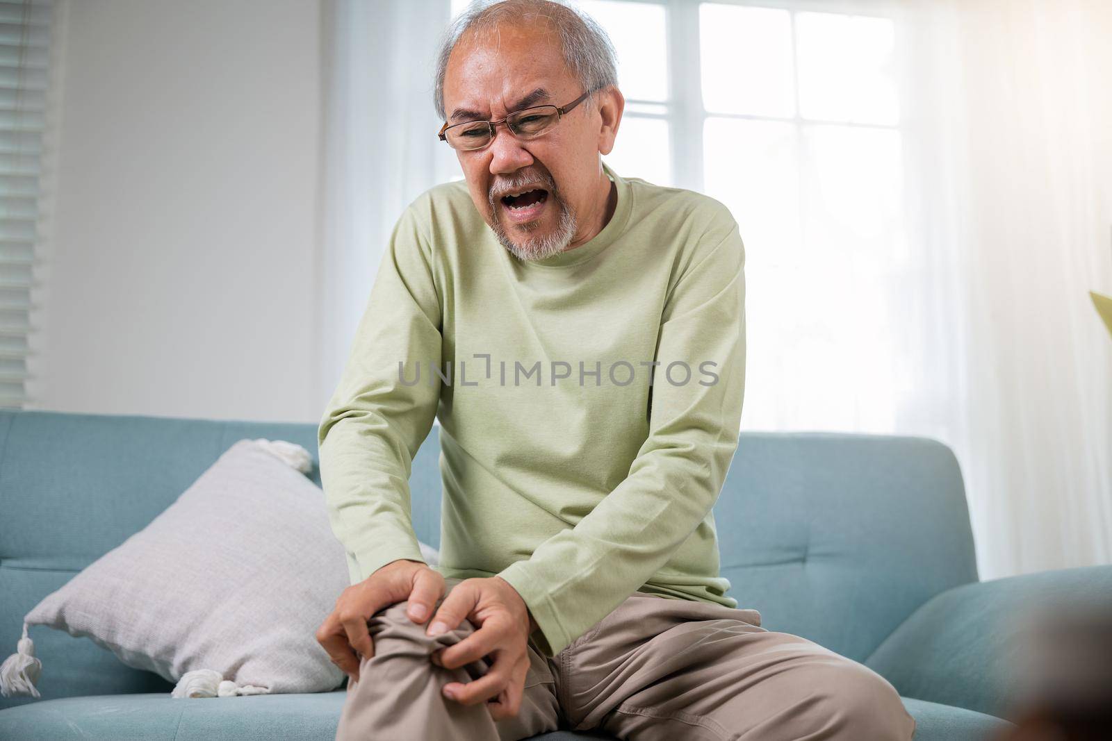 Old man suffering from knee joint pain on sofa living room, bone pain in elderly at home, senior man knee problem painful, unhappy old age hand holding on knee pain after tendon surgery