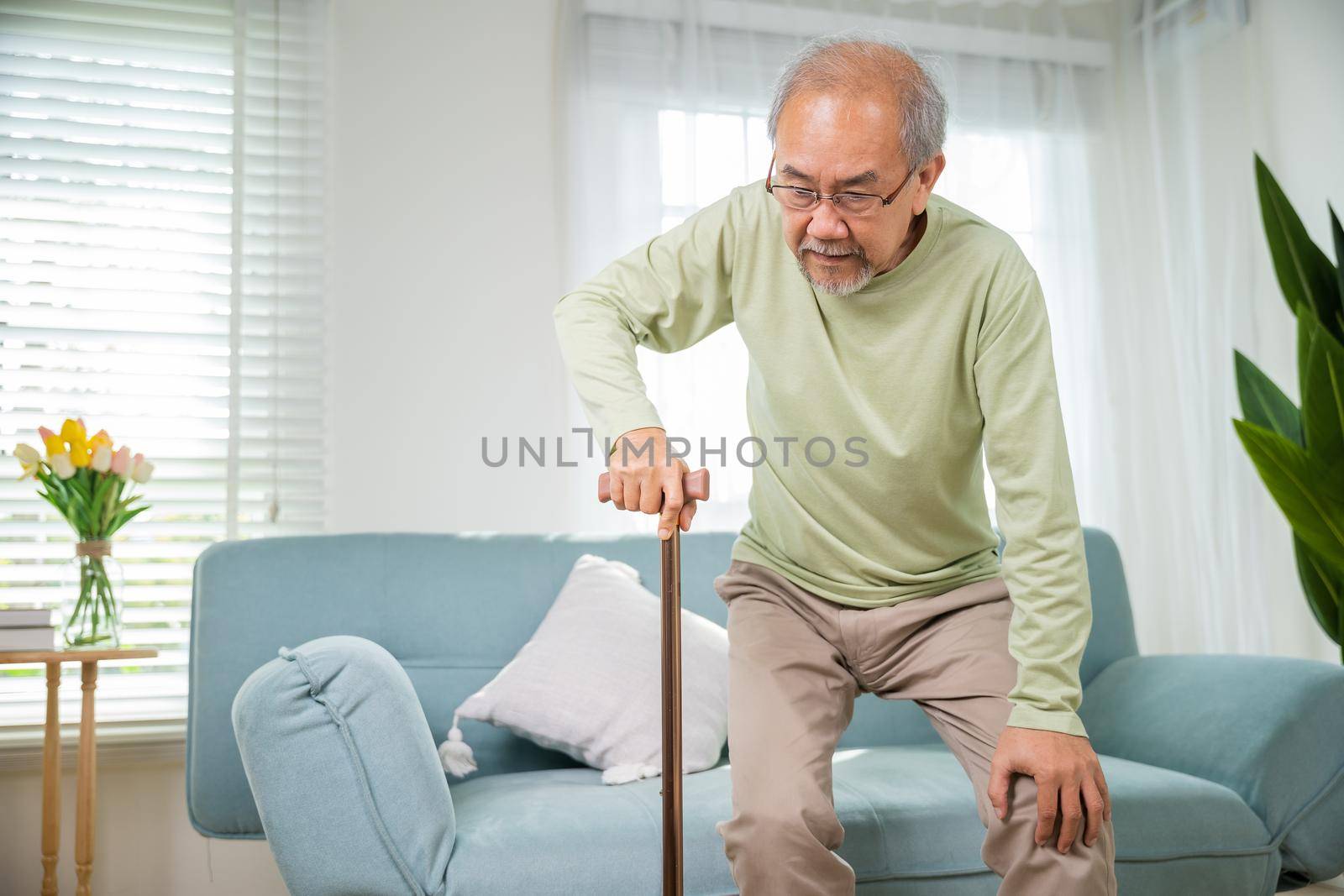 Asian senior old man with eyeglasses type to standing up from sofa with walking cane stick to walk at home, Elderly suffering from knee pain ache holding handle of cane, retirement medical healthcare