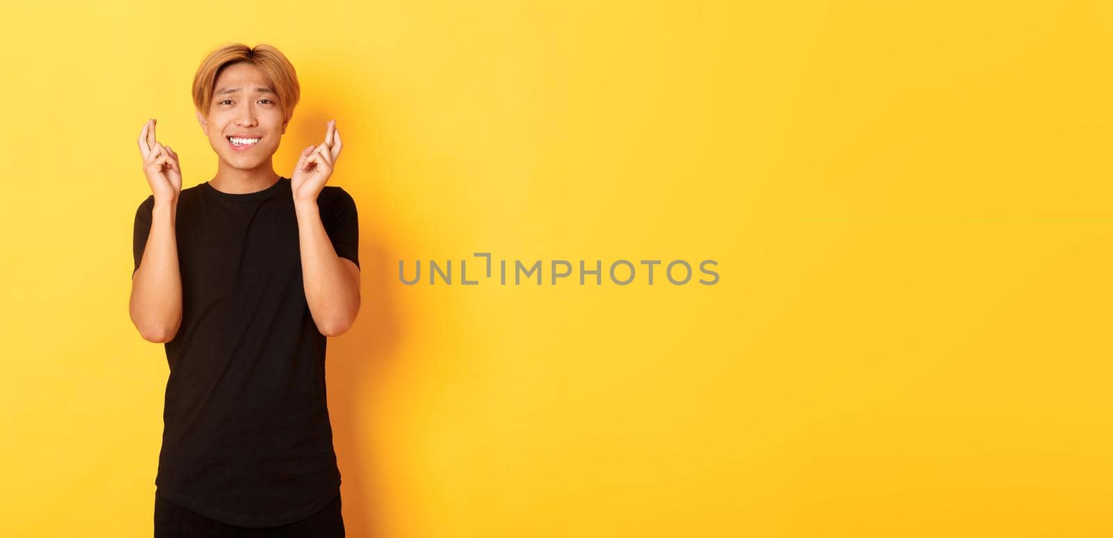 Portrait of nervous asian guy cross fingers good luck, making wish, praying over yellow background.