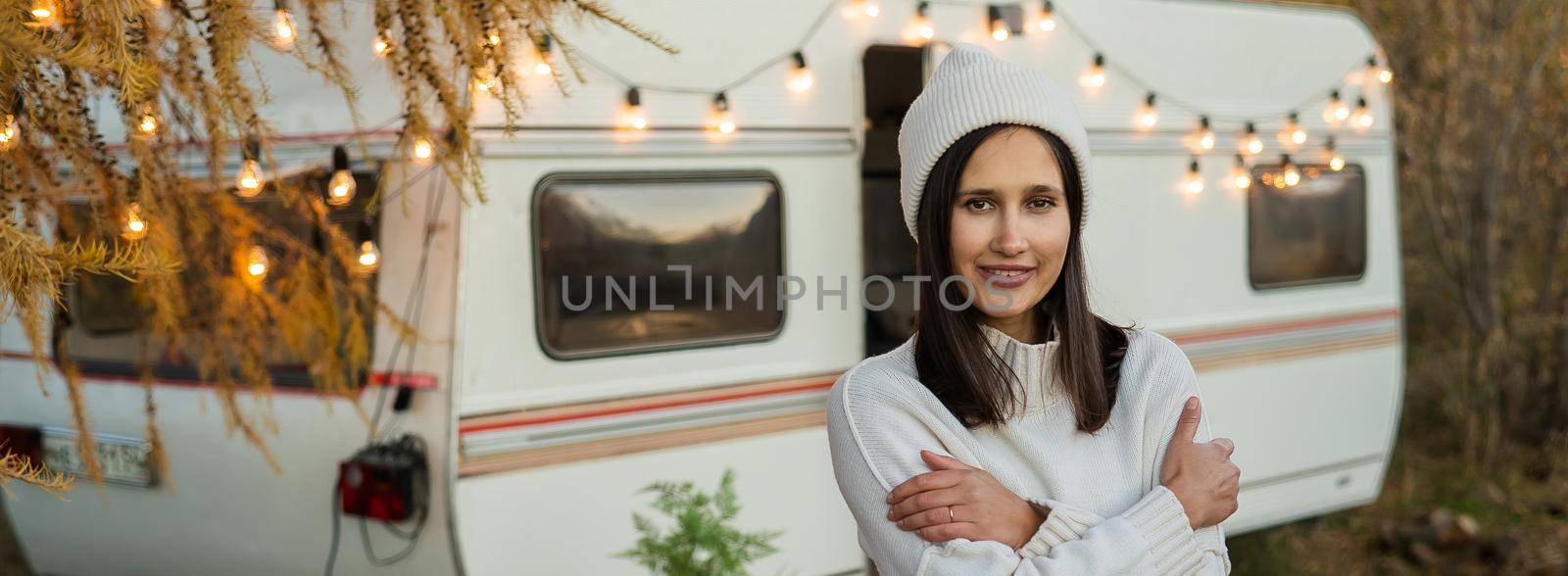 A pensive Caucasian woman in a white knitted sweater and a hat is resting near a motorhome in a warm autumn. Travel by camper