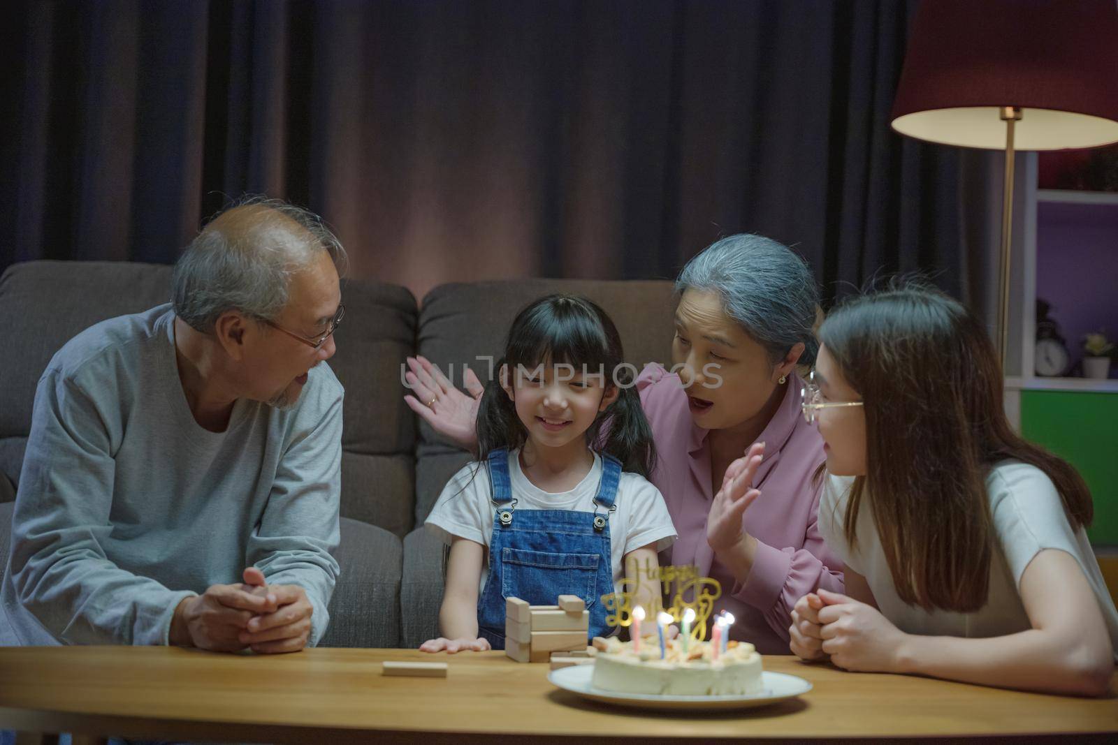 Birthday surprise. Happy Asian family grandparents and mother celebrating enjoy birthday party with cake of granddaughter at home in night time