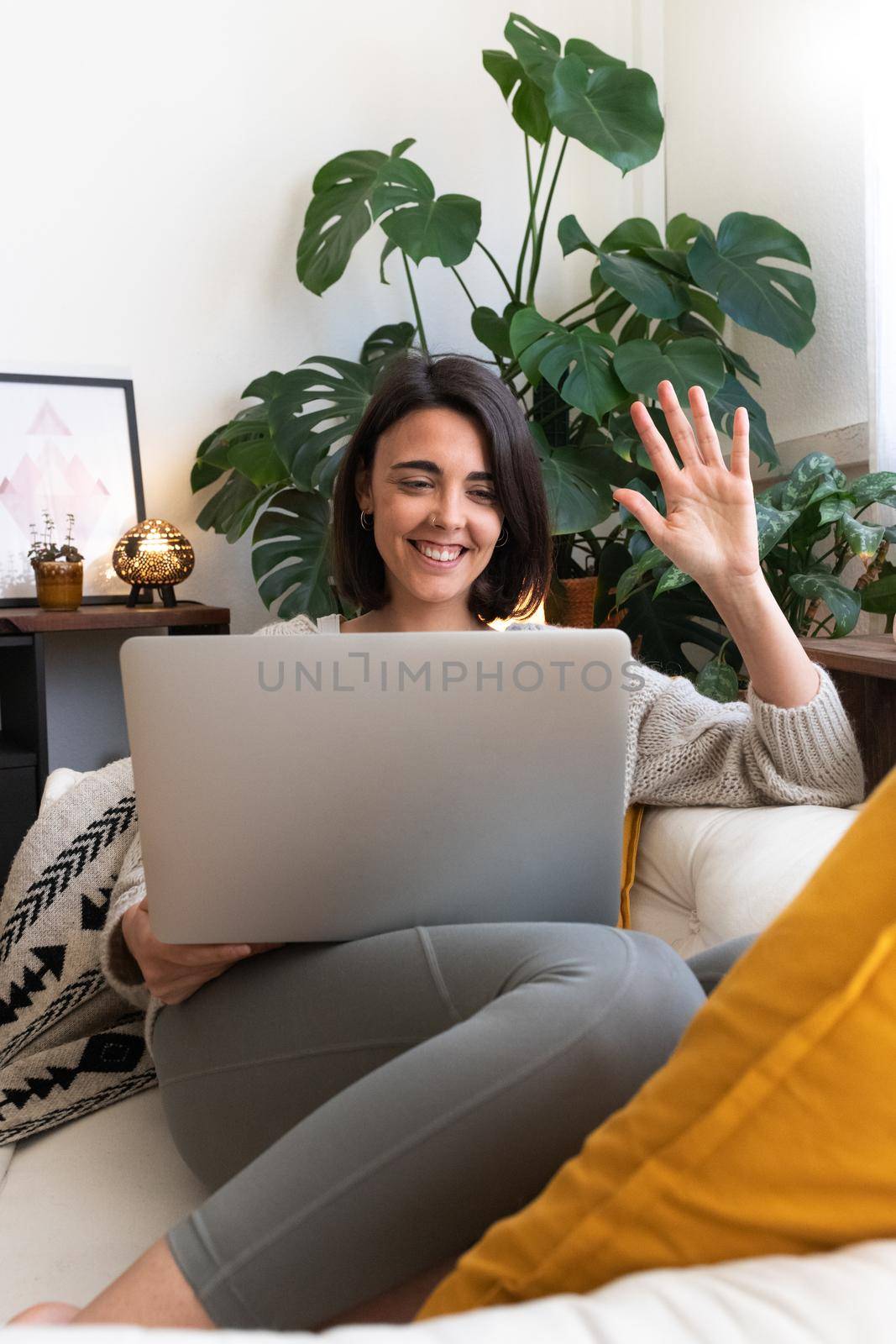 Smiling young caucasian woman lying on sofa waves hand during online video call using laptop at home. Vertical by Hoverstock