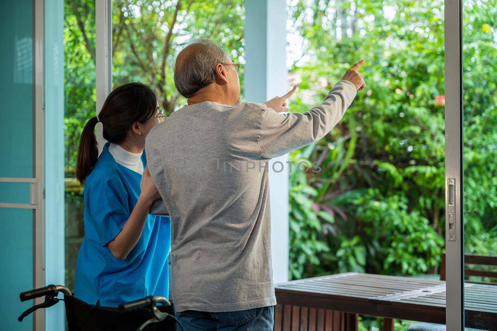 Young Asian nurse is supporting elderly man sitting in wheelchair walking to looking out window on garden and breathe fresh air, pension retirement center, senior man at home, healthcare concept