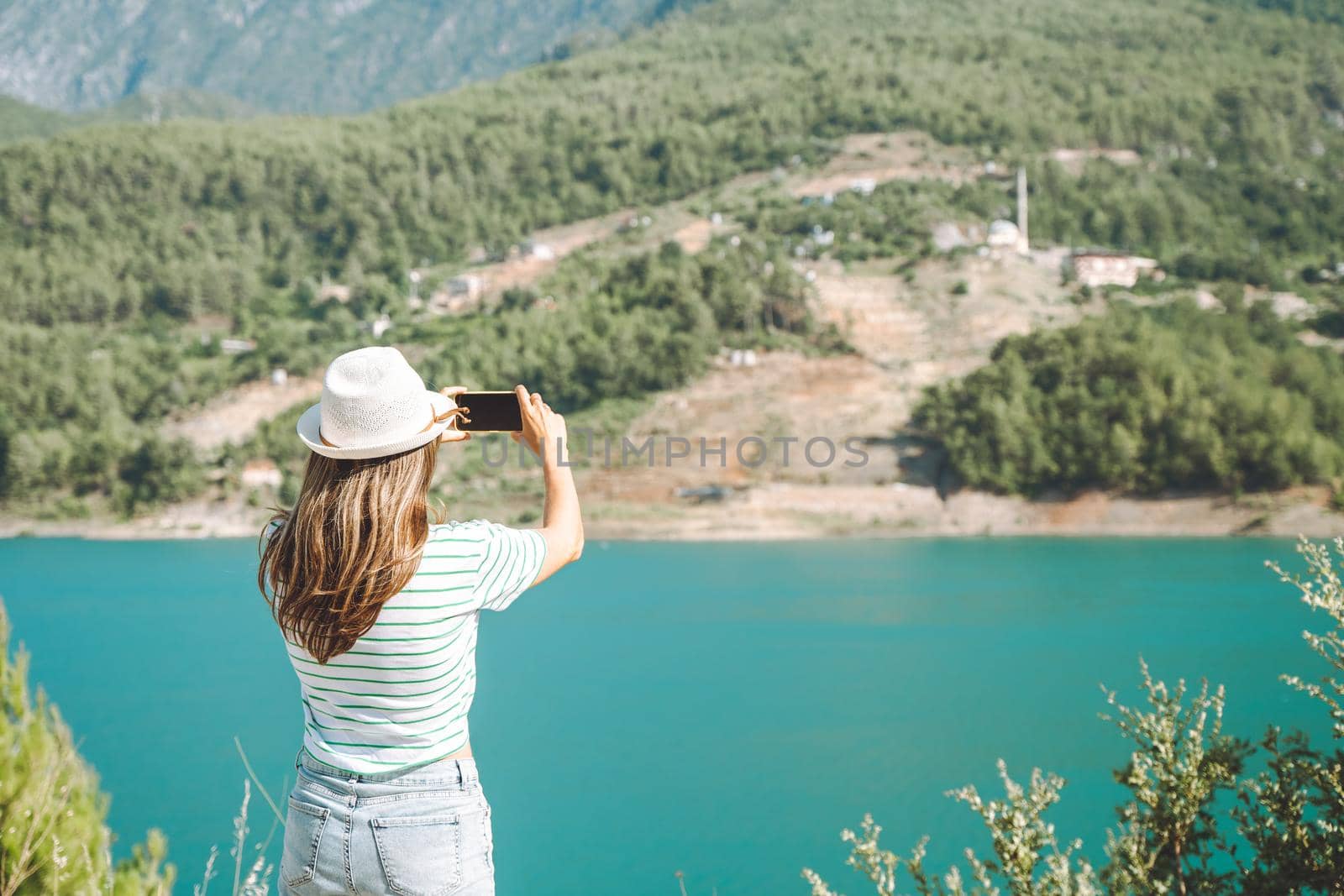 Woman in hat with wild hair shooting a video on mobile phone of mountains lake background. Traveler taking photo on cellphone on the blue lake outdoors travel adventure vacation.