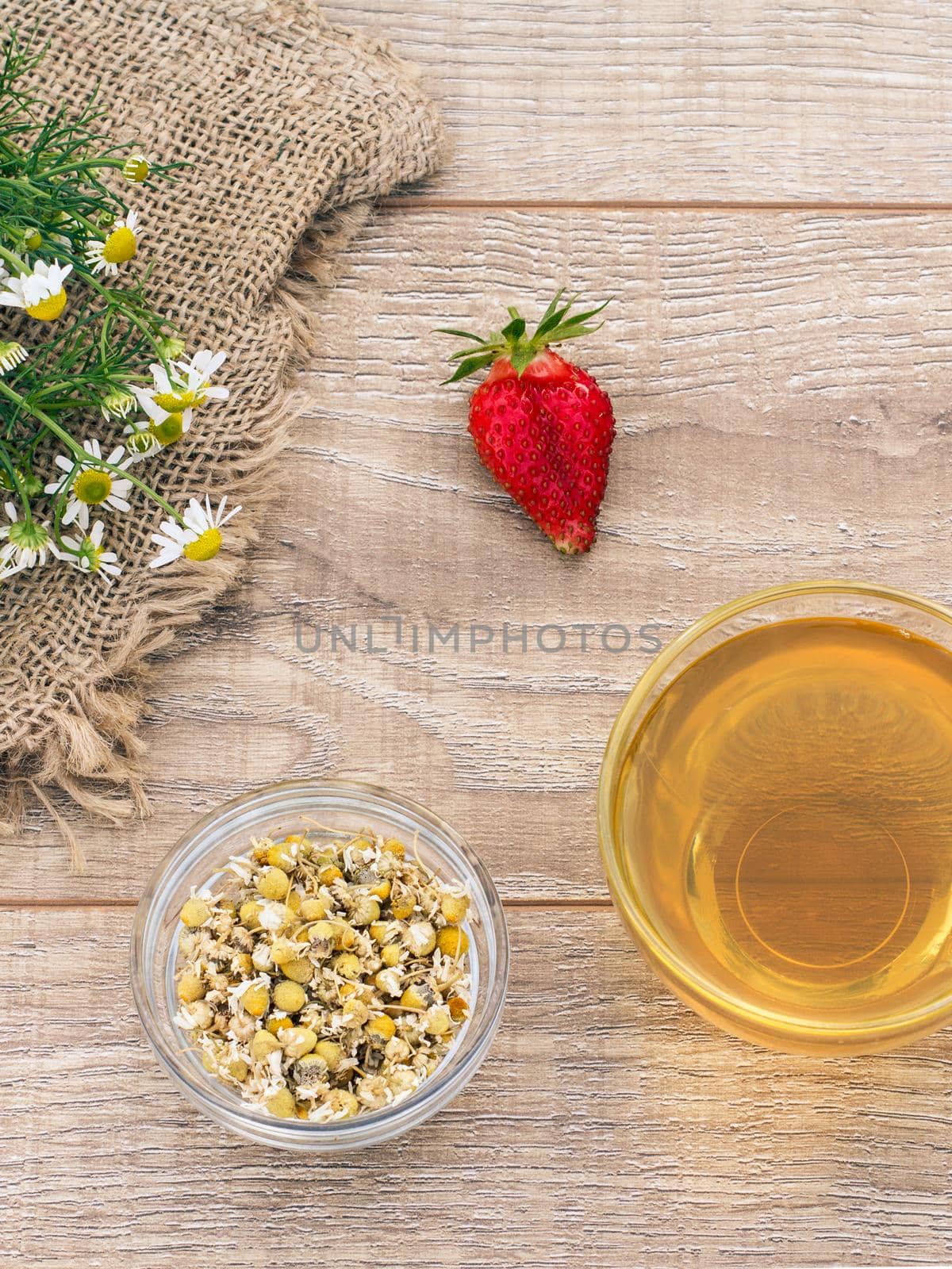 Glass cup of green tea, fresh chamomile flowers, strawberry and a little glass bowl with dry flowers of matricaria chamomilla on the wooden background. Top view.