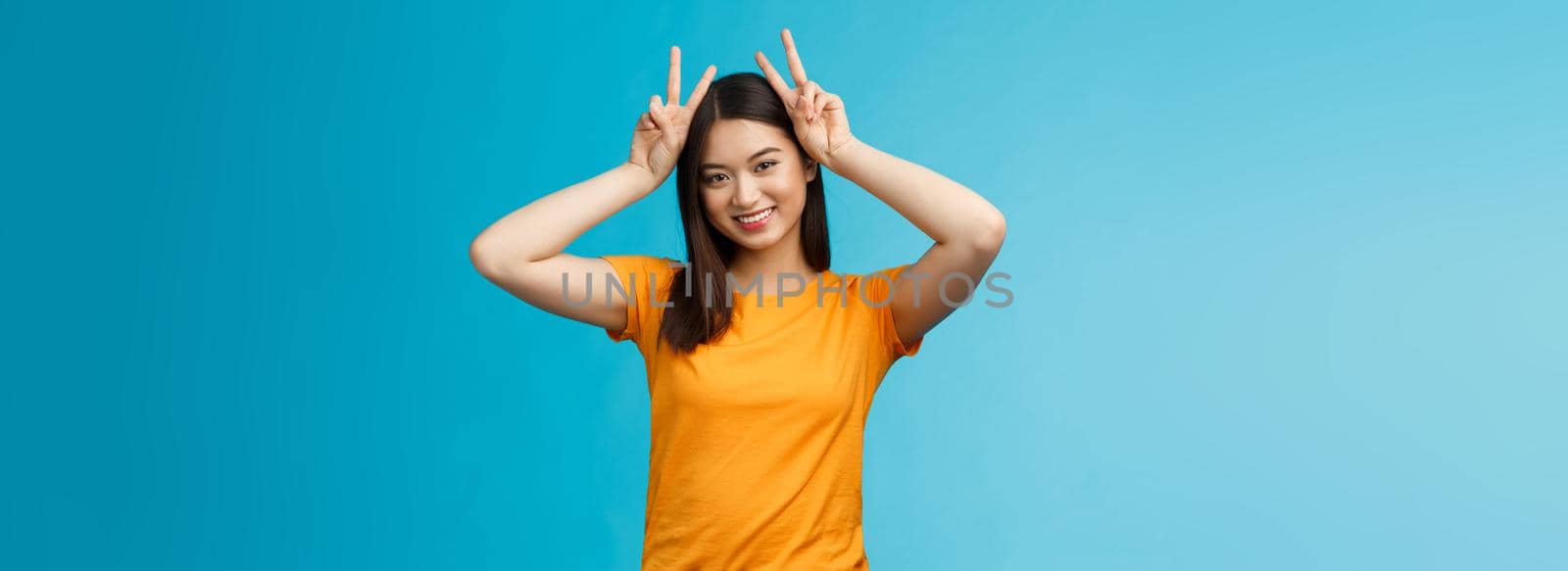 Cute tender asian girlfriend act silly lovely smile, tilt head show playfully ears hold peace victory signs on head, grinning toothy, express joy happiness and delight, stand blue background.