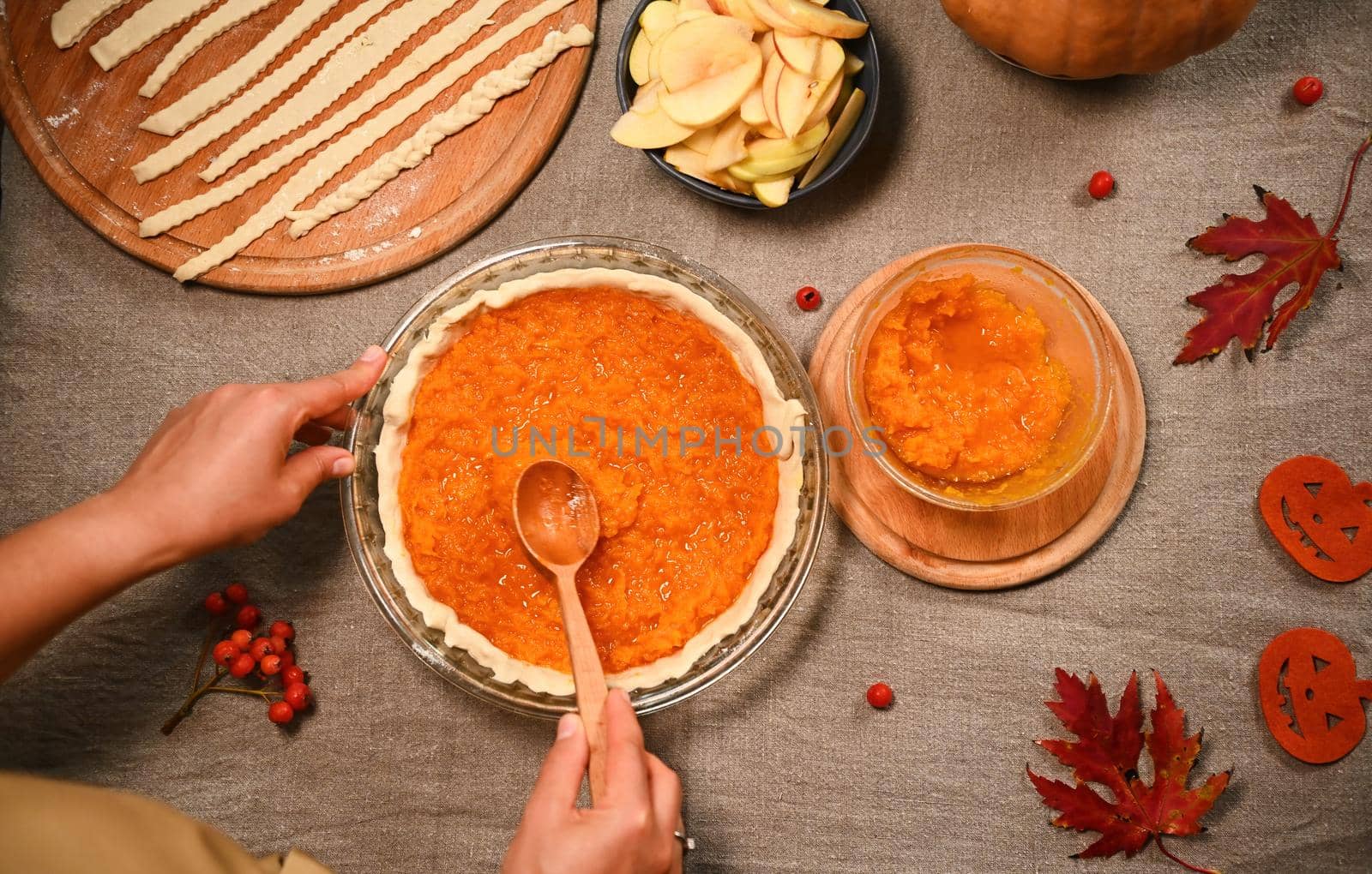 Top view making homemade American classic pumpkin pie for Thanksgiving Day. Red autumn maple leaves, viburnum berries, mashed pumpkin, apple slices and strips for making flaky crust on kitchen table