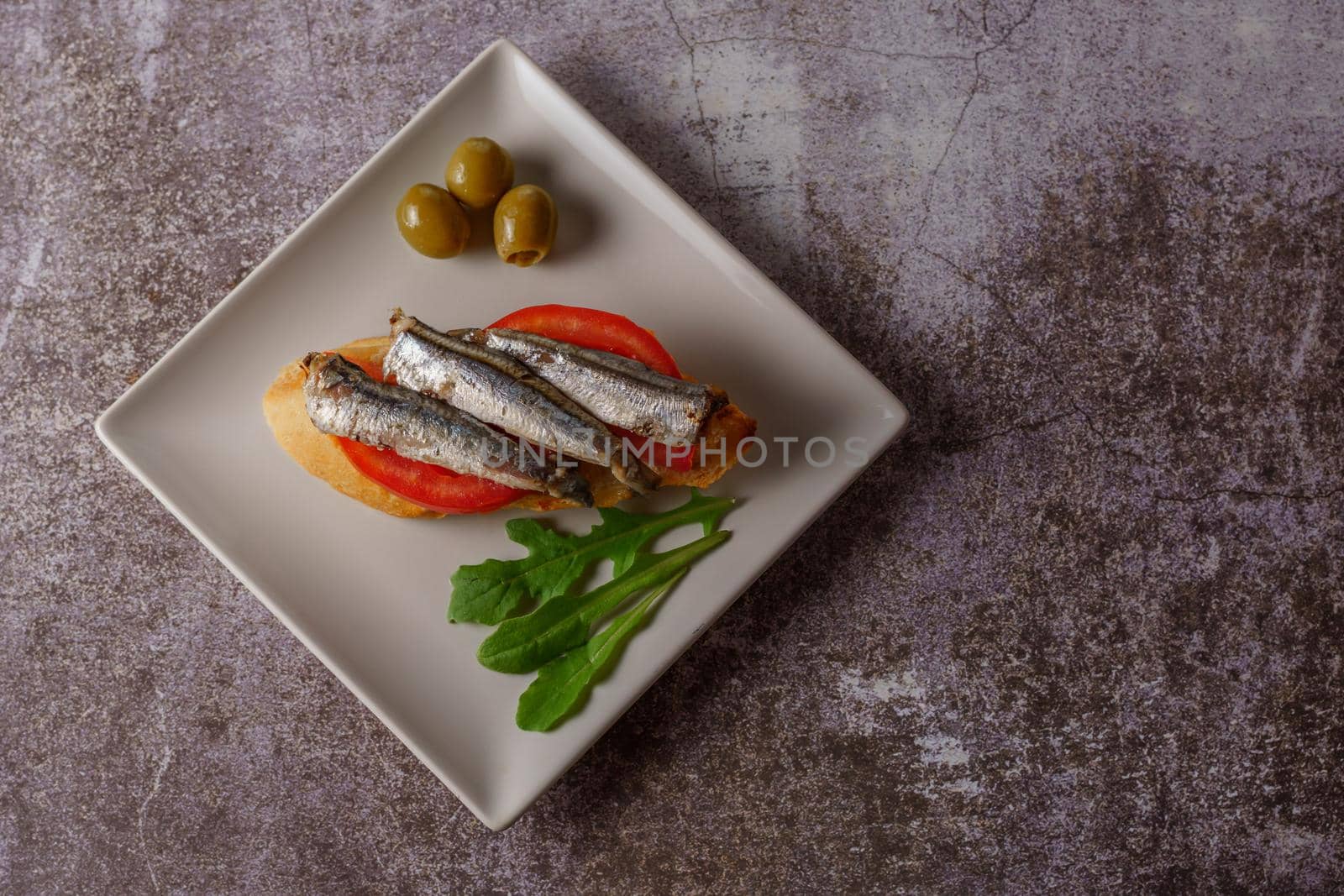 tapa of sardines on a slice of bread with tomato and olives on a white plate with a typical spanish white background