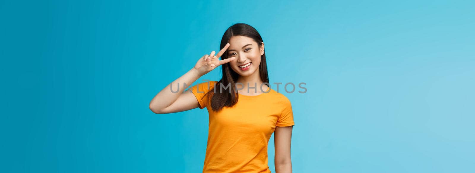 Friendly cheerful asian girlfriend look joy happiness, show peace victory sign, smiling toothy positive, enjoy lucky perfect summer sunny day, stand blue background carefree wear yellow t-shirt by Benzoix