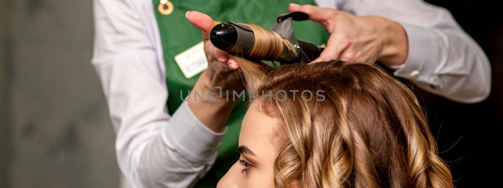 The female hairdresser is curling hair for a brown-haired young caucasian woman in a beauty salon. by okskukuruza