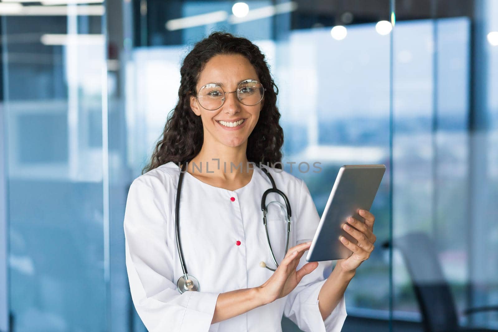 Portrait of a Latin American female doctor. Standing in the corridor of the hospital. He works, holds the tablet in his hands, uses it, types. He looks at the camera, smiles.