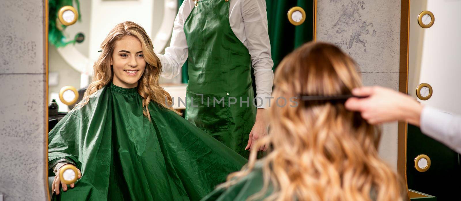 Hairdresser combing long hair of young caucasian woman looking and smiling in the mirror in beauty salon