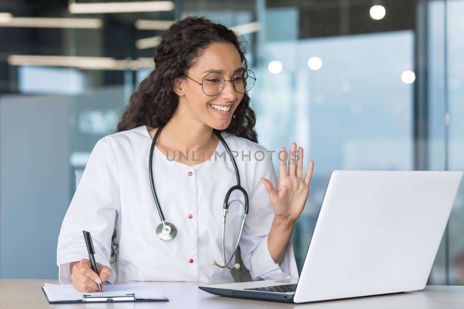 The doctor is online. A young Latin American female doctor participates in an online medical conference, meeting. He waves his hand at the laptop camera, says hello, smiles, takes notes.