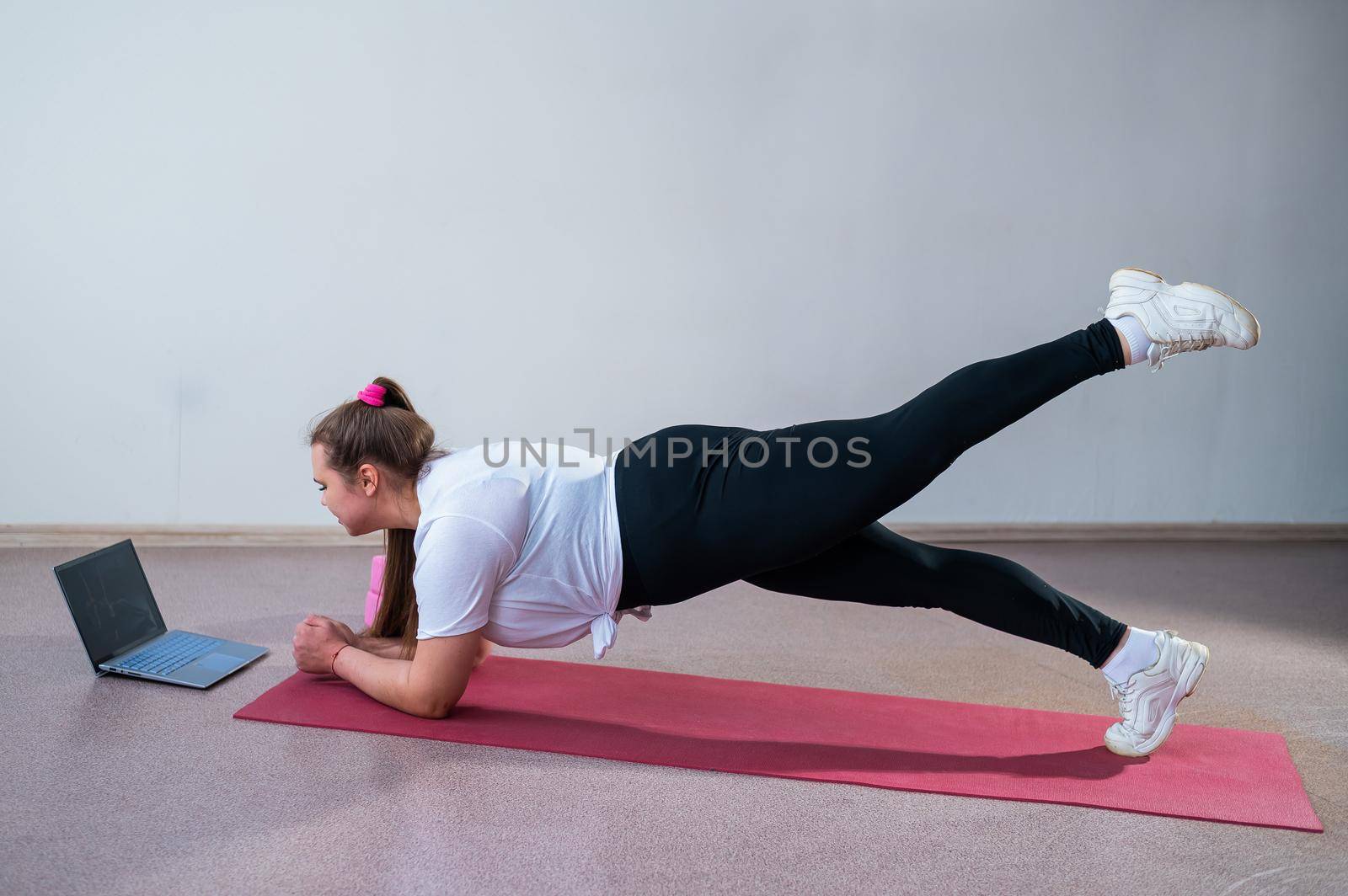 Young fat caucasian woman doing a plank on a sports mat. A cute plus size girl in sportswear is doing fitness exercises and watching an online tutorial on a laptop by mrwed54