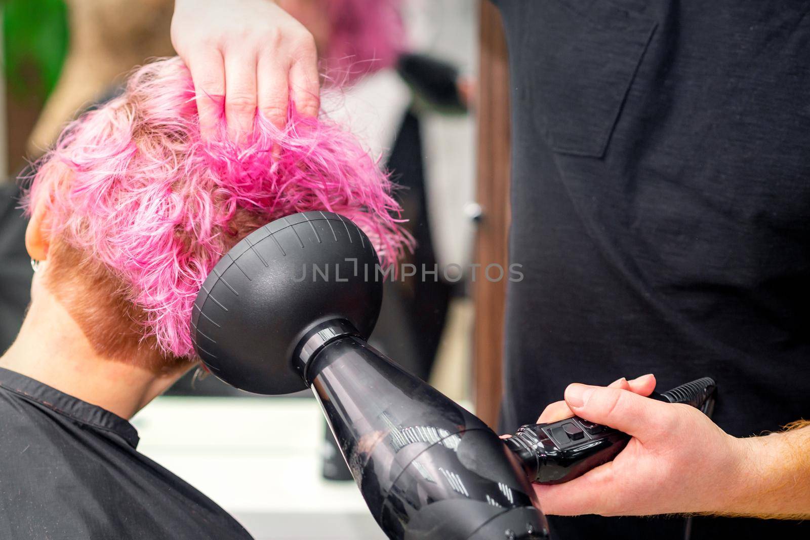 Drying short pink bob hairstyle of a young caucasian woman with a black hair dryer with the brush by hands of a male hairdresser in a hair salon, close up