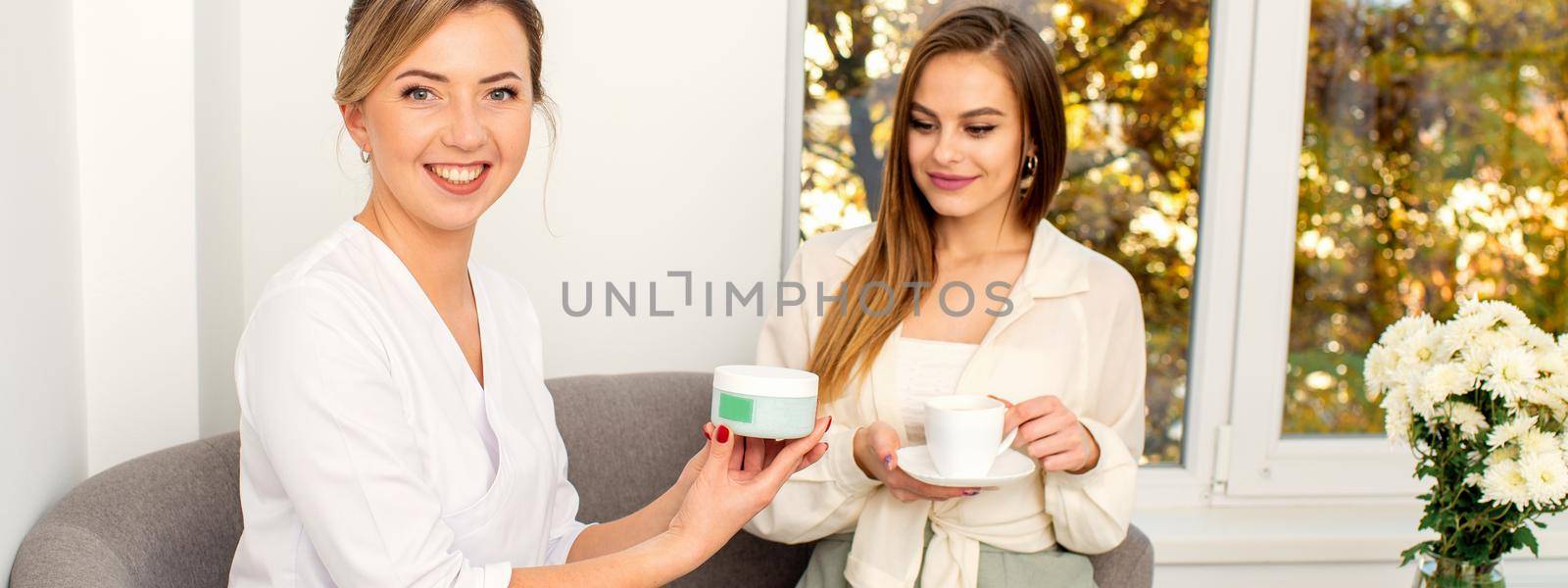 Beautician offering product for the young woman holding a white plastic jar with a cream sitting on the sofa