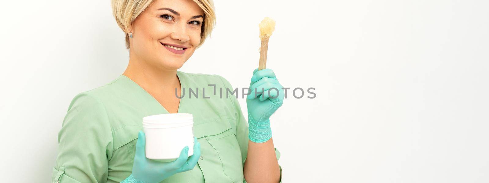 The master of sugar hair removal holds liquid yellow sugar paste, wax for depilation with the jar on a wooden stick on a white background. by okskukuruza