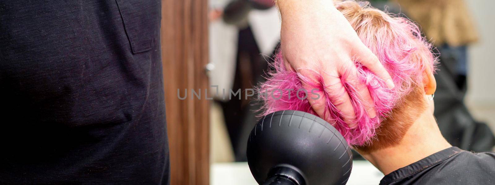 Drying short pink bob hairstyle of a young caucasian woman with a black hair dryer with the brush by hands of a male hairdresser in a hair salon, close up