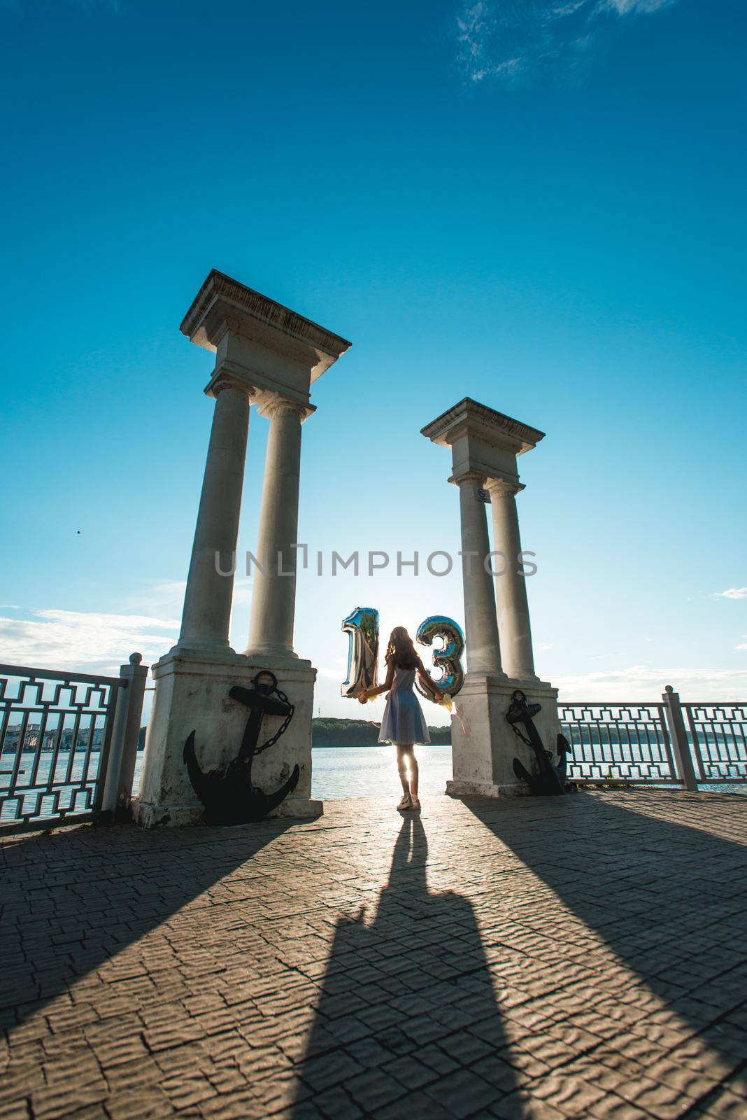 Happy teenage party girl 12-13 years old with balloons. Standing on the lake side between columns. look from the back. silhouette