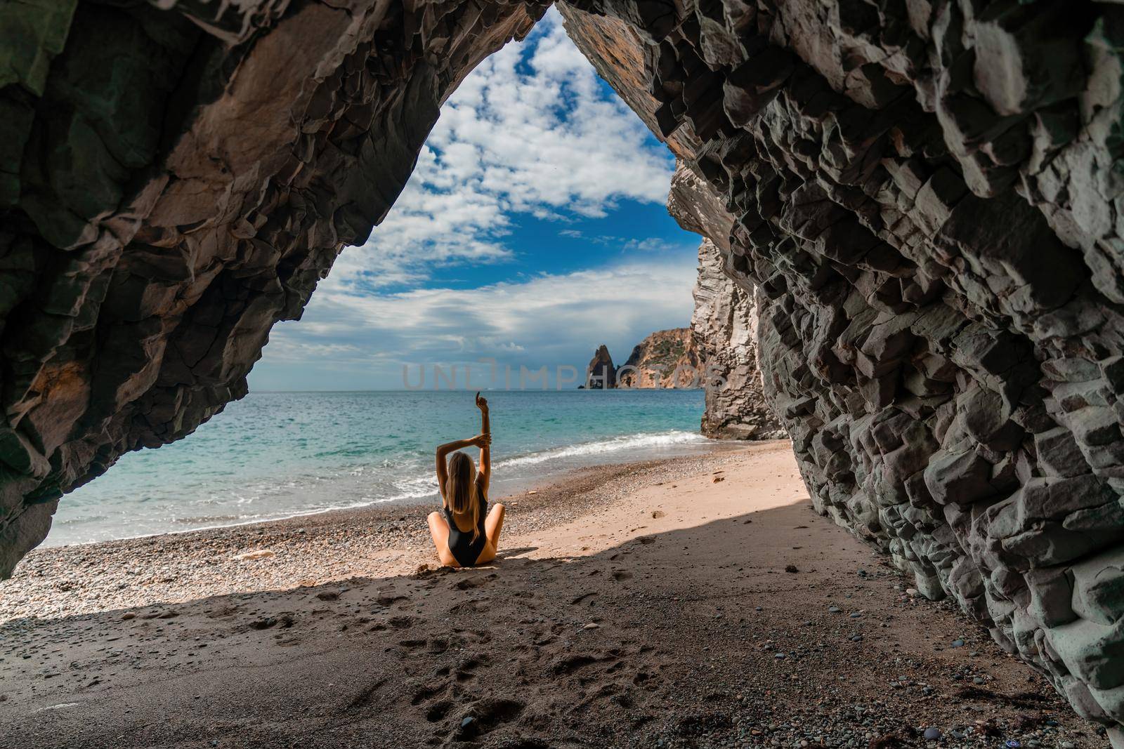 View of a woman in a black swimsuit from a sea cave Attractive woman enjoying the sea air sits on the beach and looks at the sea. Behind her are rocks and the sea by Matiunina