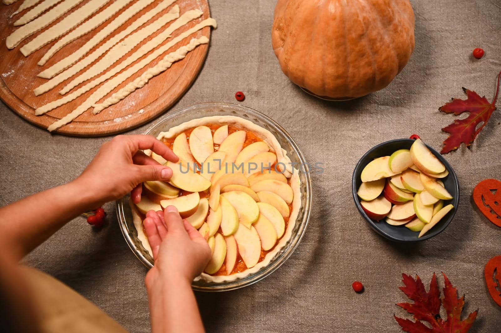 Top view of housewife, chef confectioner putting slices of apples on a raw pumpkin pie, preparing homemade traditional American classic tarte with crust lattice, for Thanksgiving Day. Autumn holidays