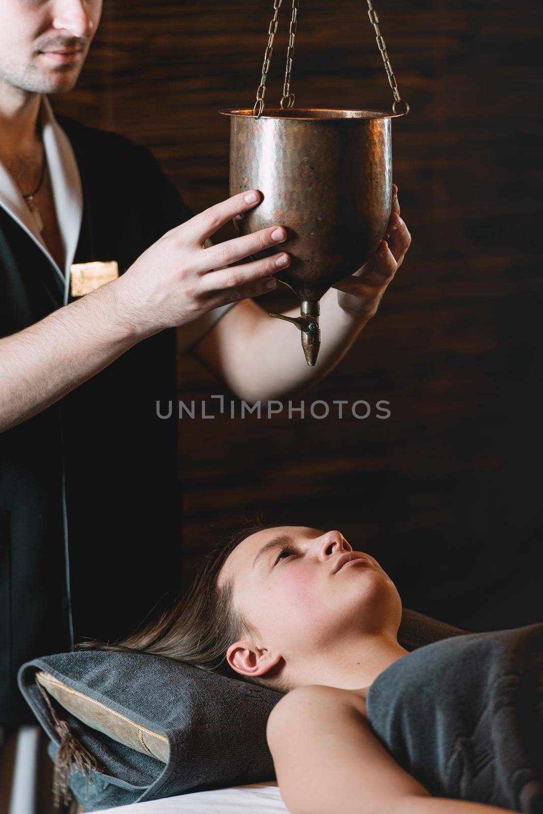 Specialist making shirodhara massage form of ayurveda therapy. Young charming girl on a panchakarma procedure laying on a massage table. beautiful woman spending time at modern spa cabinet relaxing. Soft yellow light.