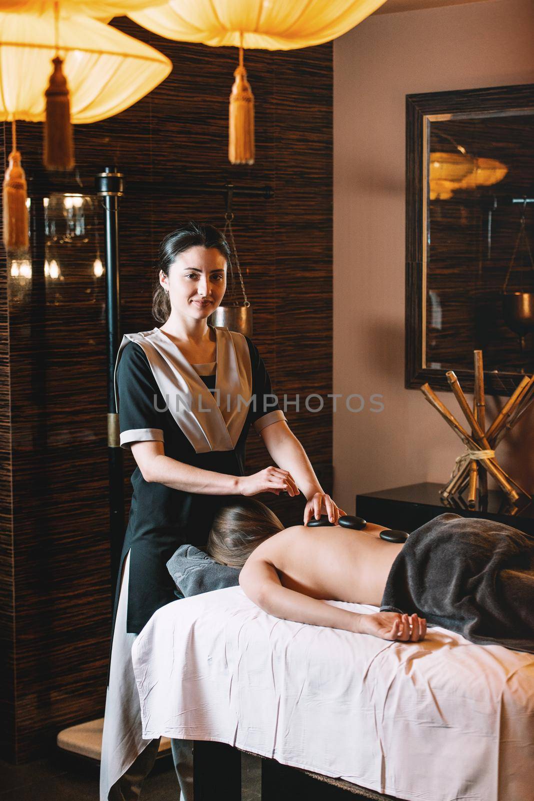 Specialist making hot stone massage to a client. Young charming girl on a panchakarma procedure laying on a massage table. beautiful woman spending time at modern spa cabinet relaxing. Soft yellow light.