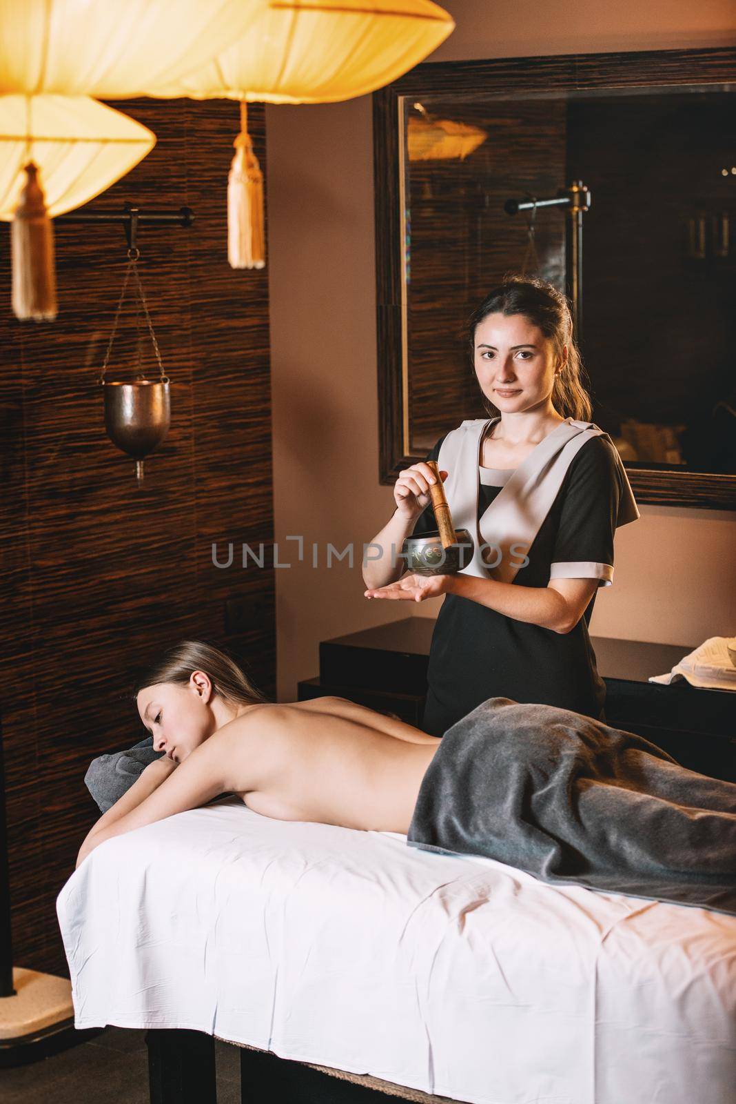 Specialist holding in hands Brass Hammer Singing Bowl. Young charming girl on a panchakarma procedure laying on a massage table. beautiful woman spending time at modern spa cabinet relaxing. Soft yellow light.