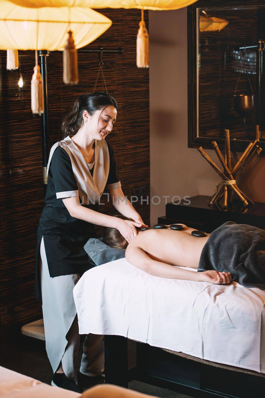 Specialist making hot stone massage to a client. Young charming girl on a panchakarma procedure laying on a massage table. beautiful woman spending time at modern spa cabinet relaxing. Soft yellow light.