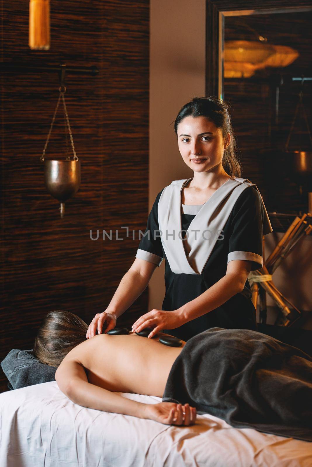 Specialist making hot stone massage to a client. Young charming girl on a panchakarma procedure laying on a massage table. beautiful woman spending time at modern spa cabinet relaxing. Soft yellow light.