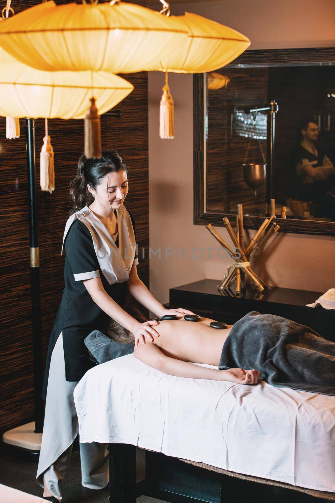 Specialist making hot stone massage to a client. Young charming girl on a panchakarma procedure laying on a massage table. beautiful woman spending time at modern spa cabinet relaxing. Soft yellow light.