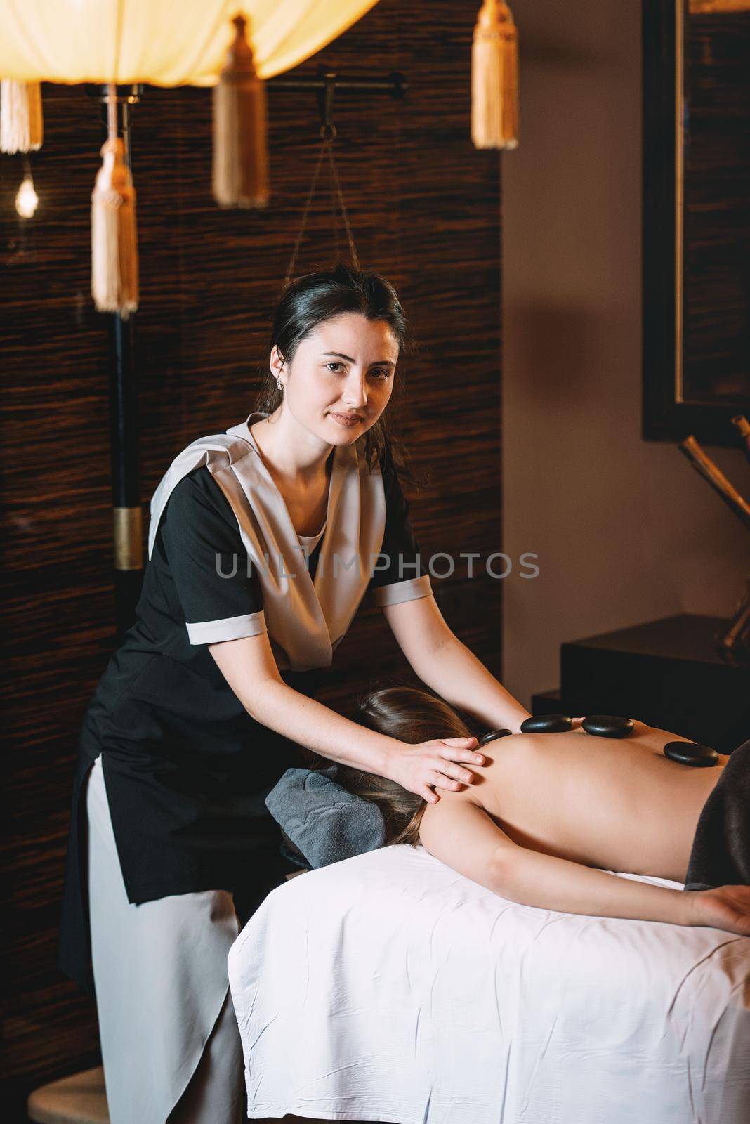 Specialist making hot stone massage to a client. Young charming girl on a panchakarma procedure laying on a massage table. beautiful woman spending time at modern spa cabinet relaxing. Soft yellow light.