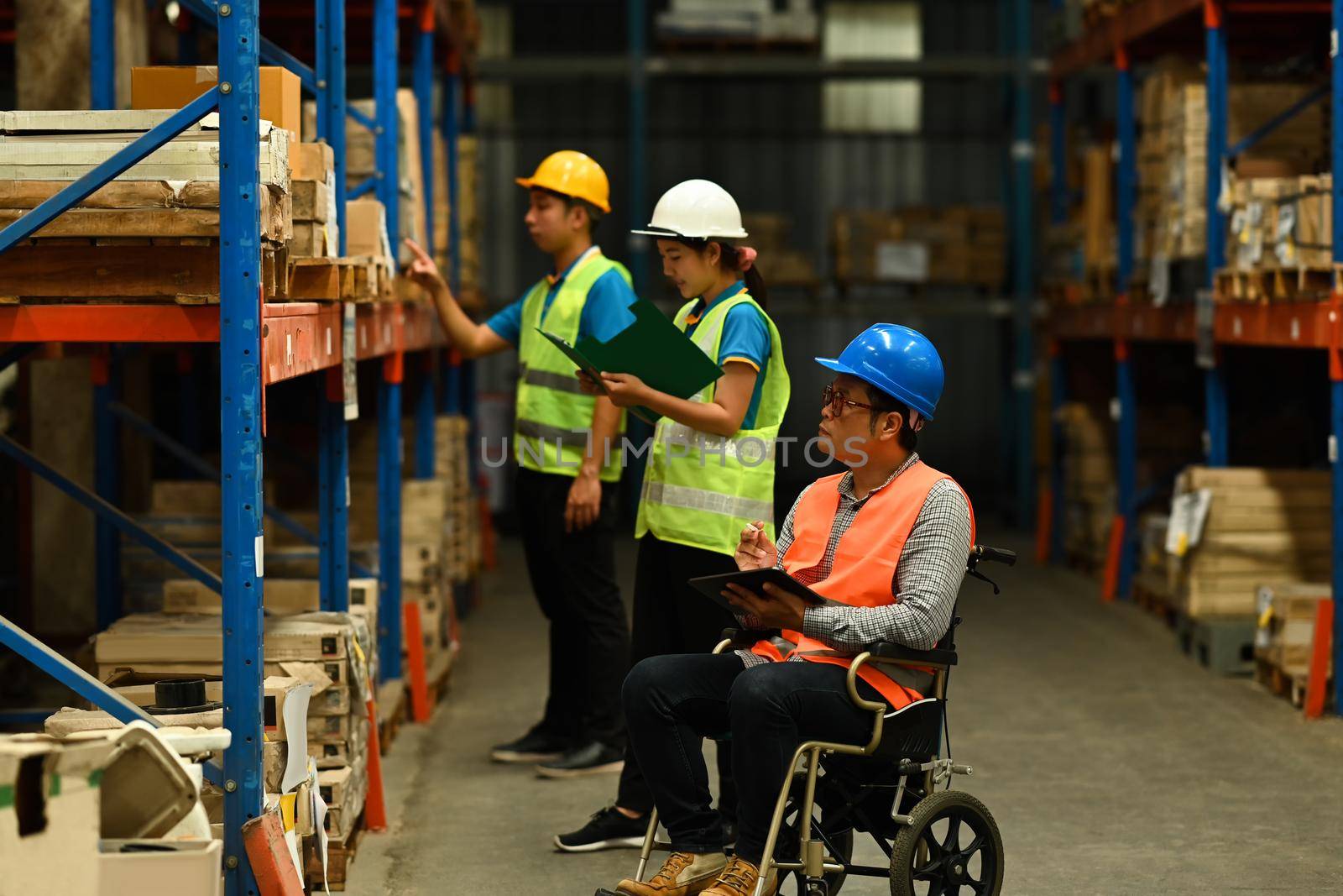 Senior male manager in wheelchair and young workers wearing safety uniform checking quantity of storage product on shelf.