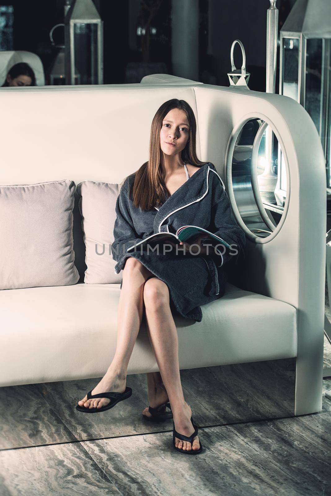Portrait of young beautiful woman relaxing in a chair waiting for a procedure. in a bathrobe with magazine in hands. Luxery spa center.