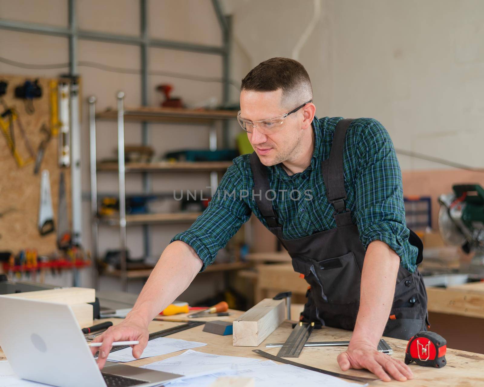 Portrait of a carpenter in protective glasses and work overalls uses a laptop in a workshop. by mrwed54