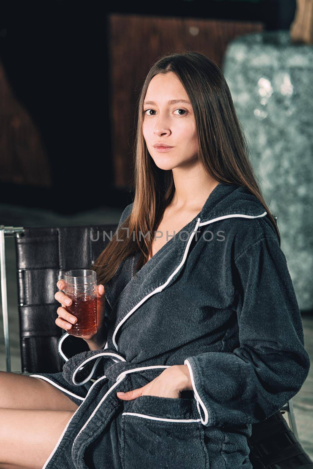 Portrait of young beautiful woman relaxing in a chair in a bathrobe with a detox drink in a hand. Luxery spa center. look at the camera