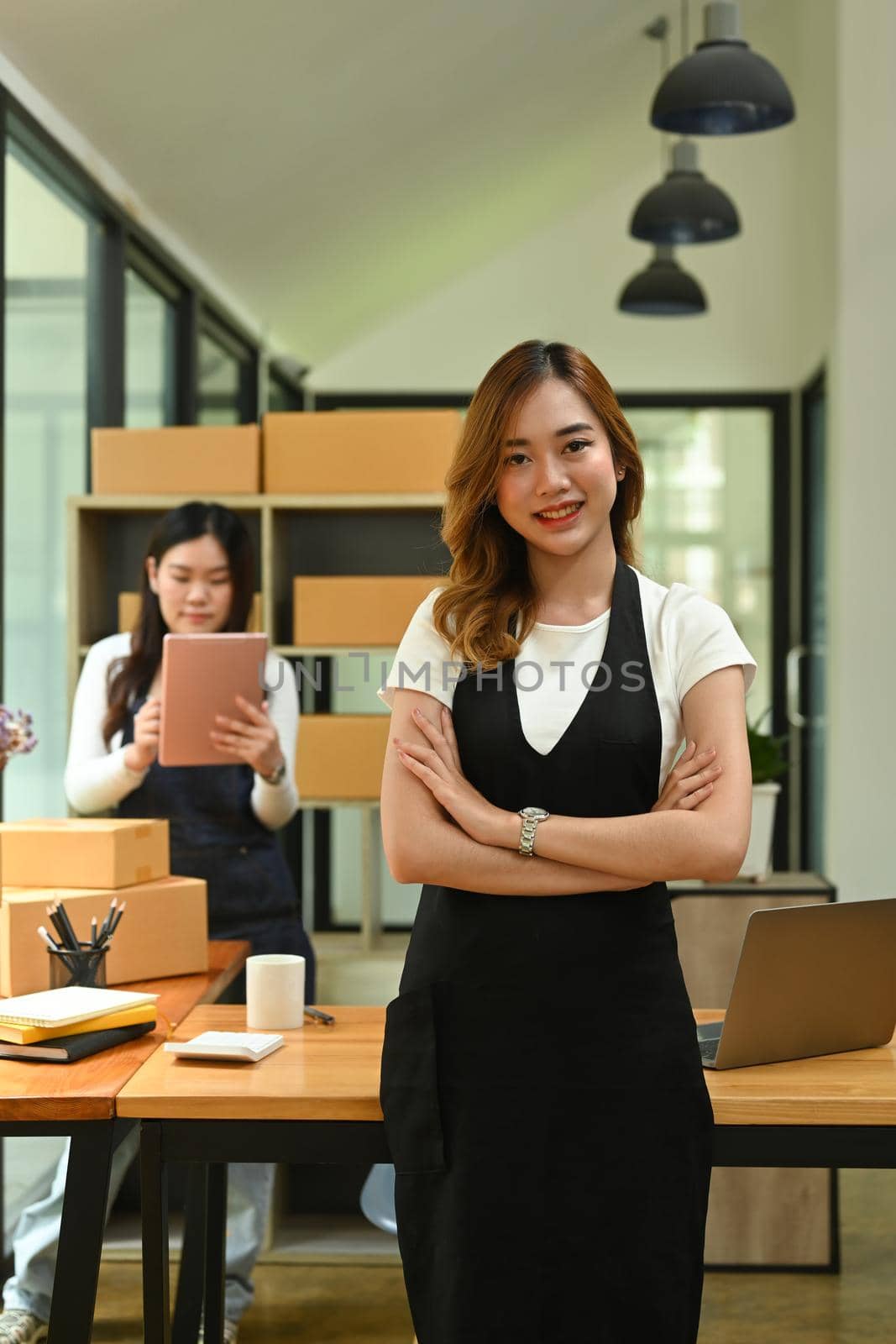 Portrait of small business owner in apron standing with arms crossed in modern home office.