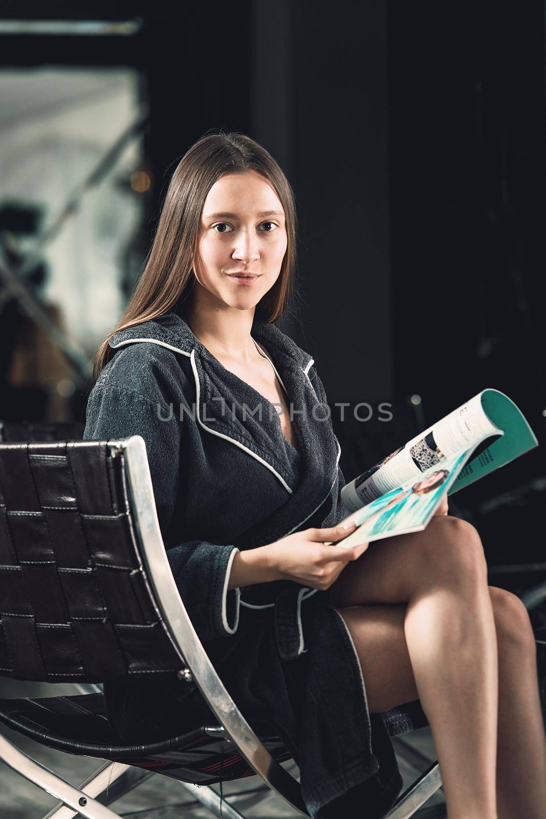 Portrait of young beautiful woman relaxing in a chair waiting for a procedure. in a bathrobe with magazine in hands. Luxery spa center.