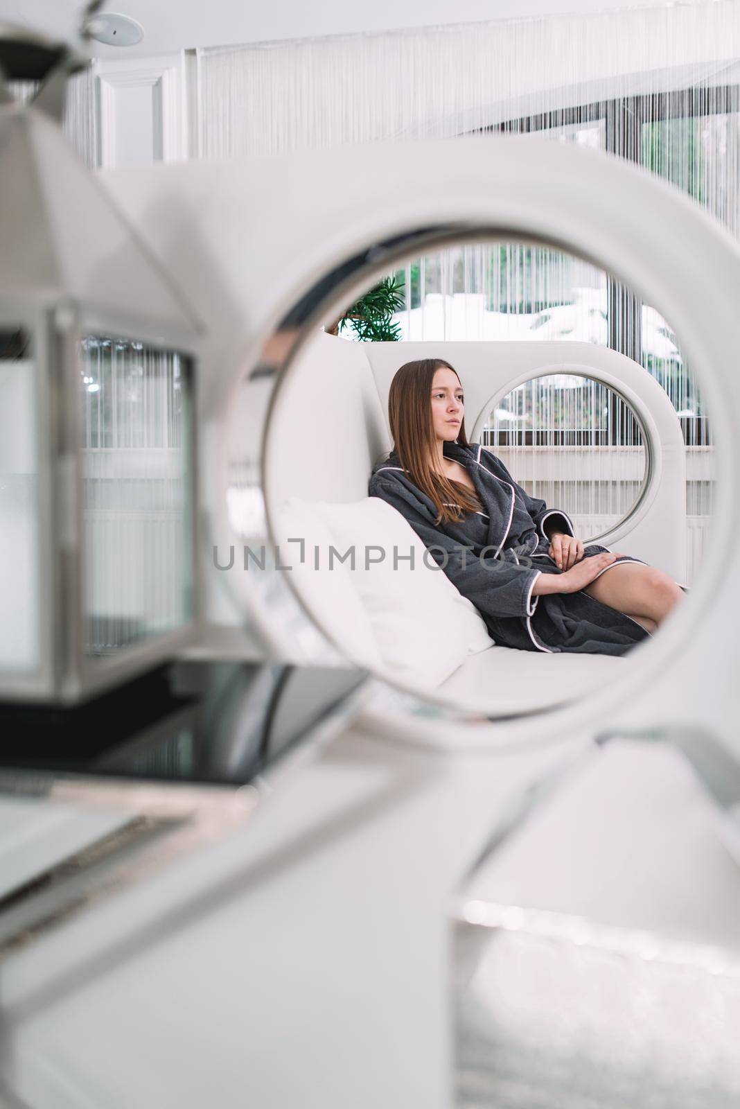 Portrait of young beautiful woman relaxing in a fashionable white sofa in a gray bathrobe, waiting for treatment. Luxery spa center