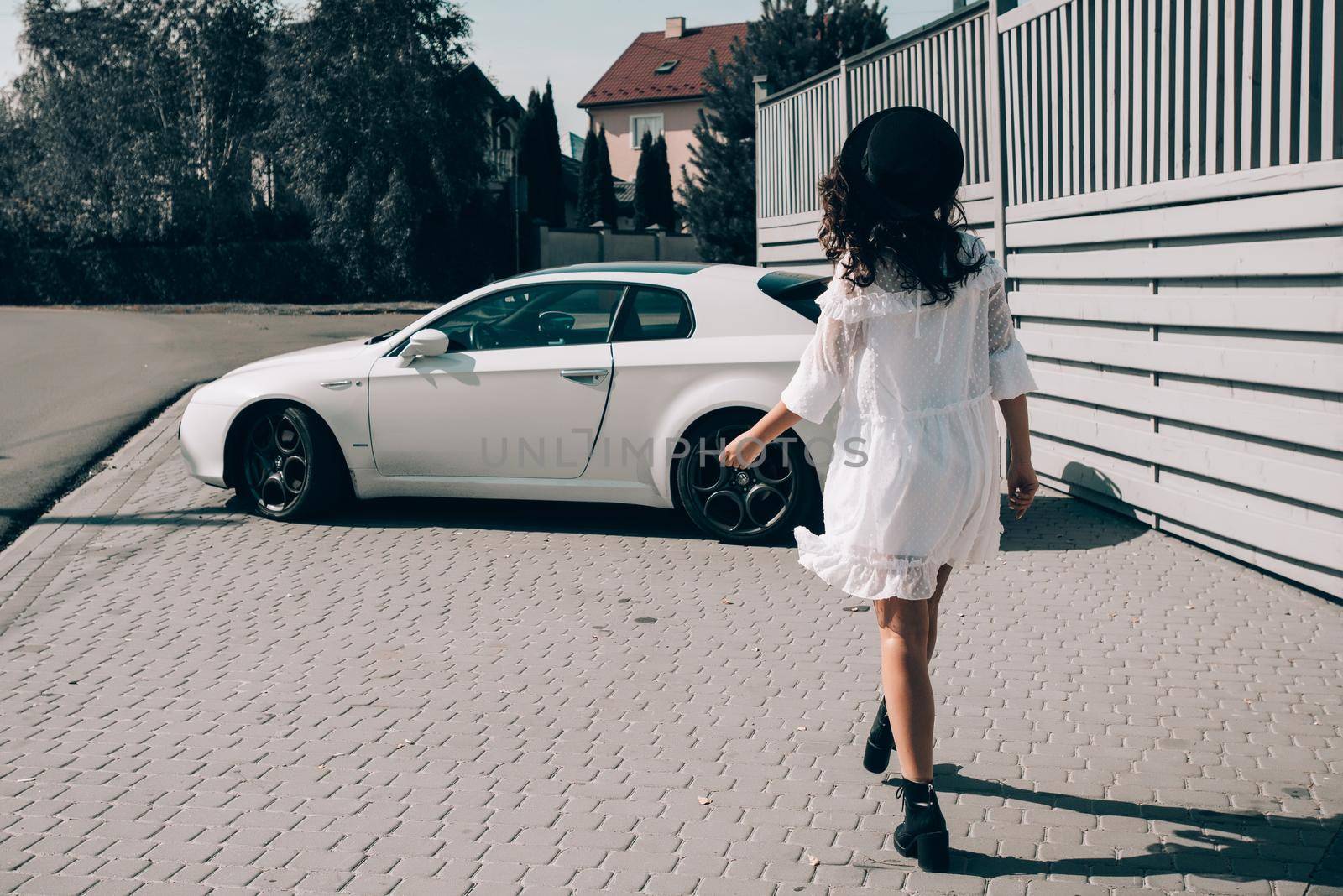 Sunny lifestyle fashion portrait of young stylish hipster woman walking to her car, wearing trendy white dress, black hat and boots. Gray wooden backgrond.