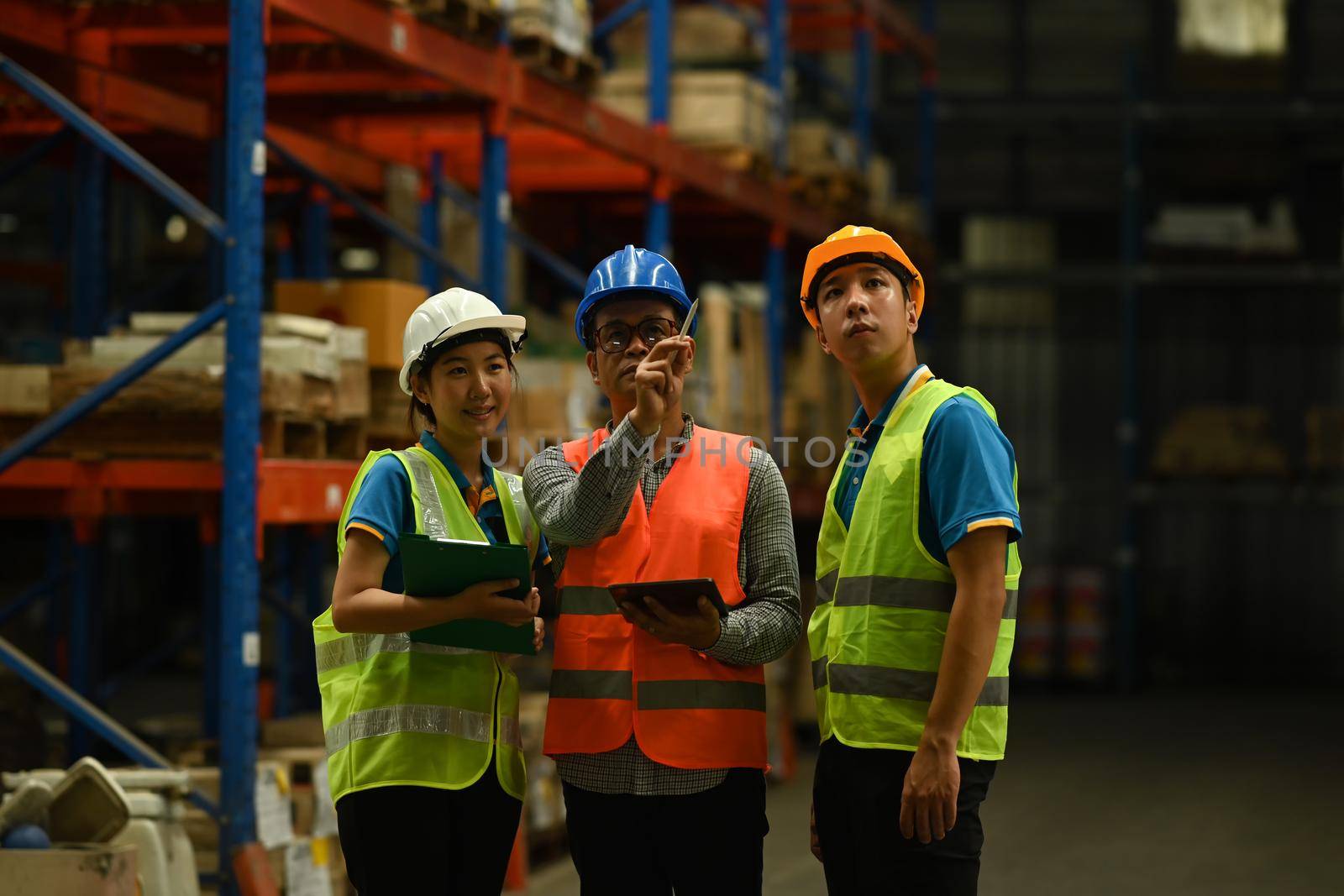 Warehouse workers and middle age manager standing between rows of tall shelves and analyzing newly arrived goods on tablet.