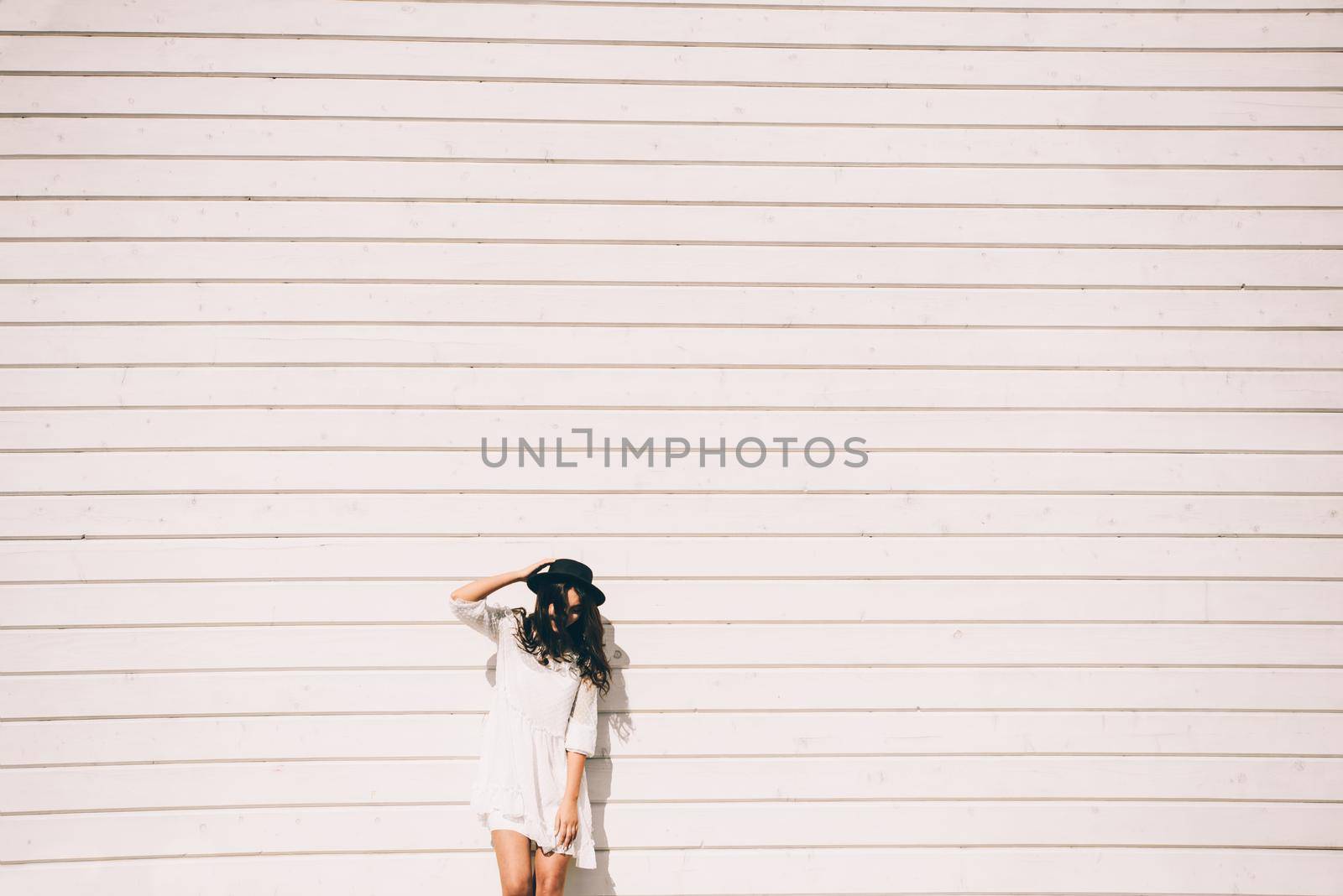 Sunny lifestyle fashion portrait of young stylish hipster woman walking on the street, wearing trendy white dress, black hat and boots. White wooden backgrond.