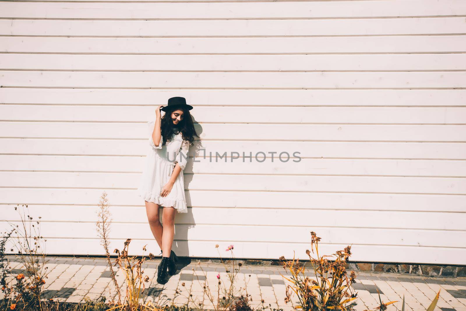 Sunny lifestyle fashion portrait of young stylish hipster woman walking on the street, wearing trendy white dress, black hat and boots. White wooden backgrond.