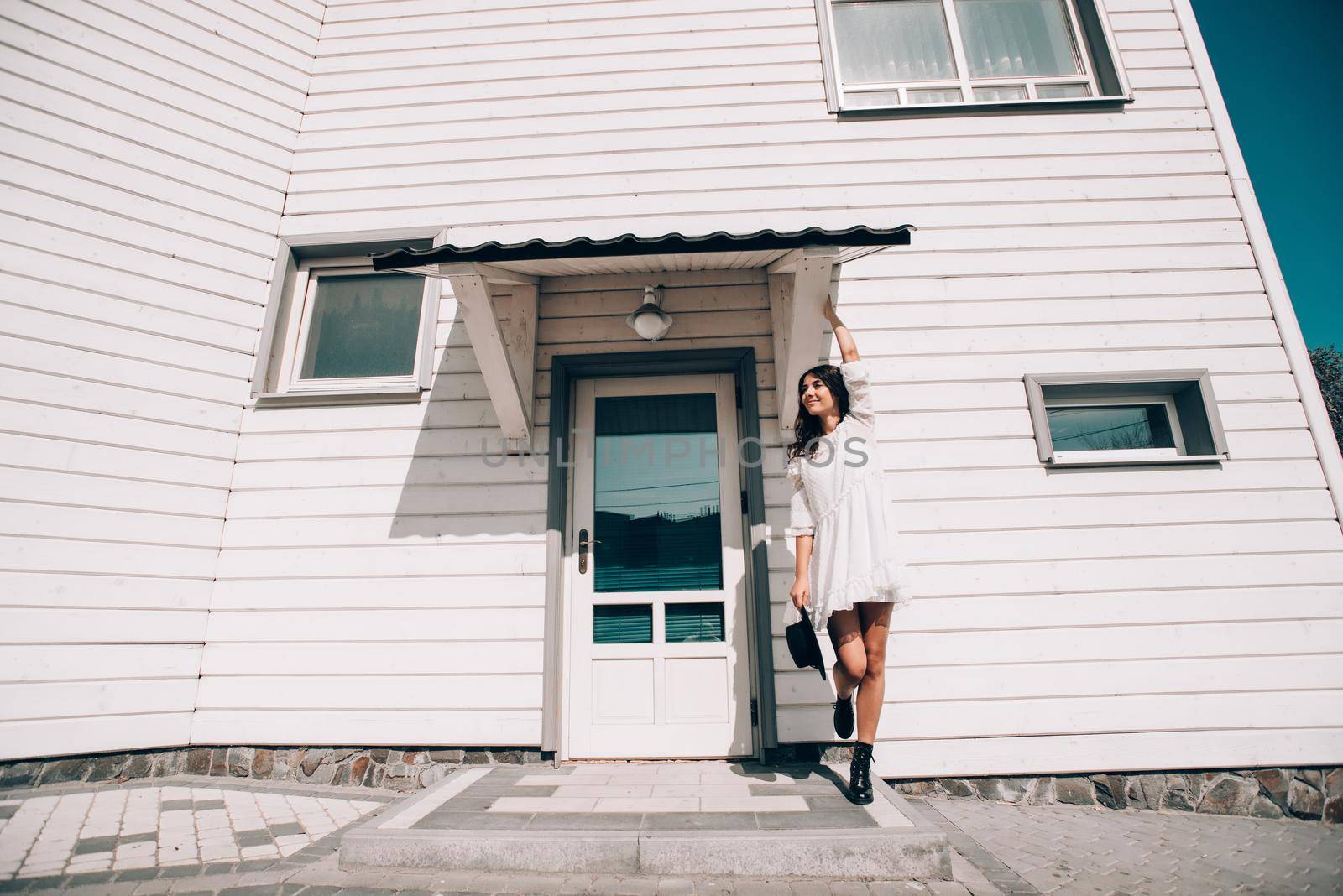 Sunny lifestyle fashion portrait of young stylish hipster woman walking on the street, wearing trendy white dress, black hat and boots. White wooden house on a backgrond.